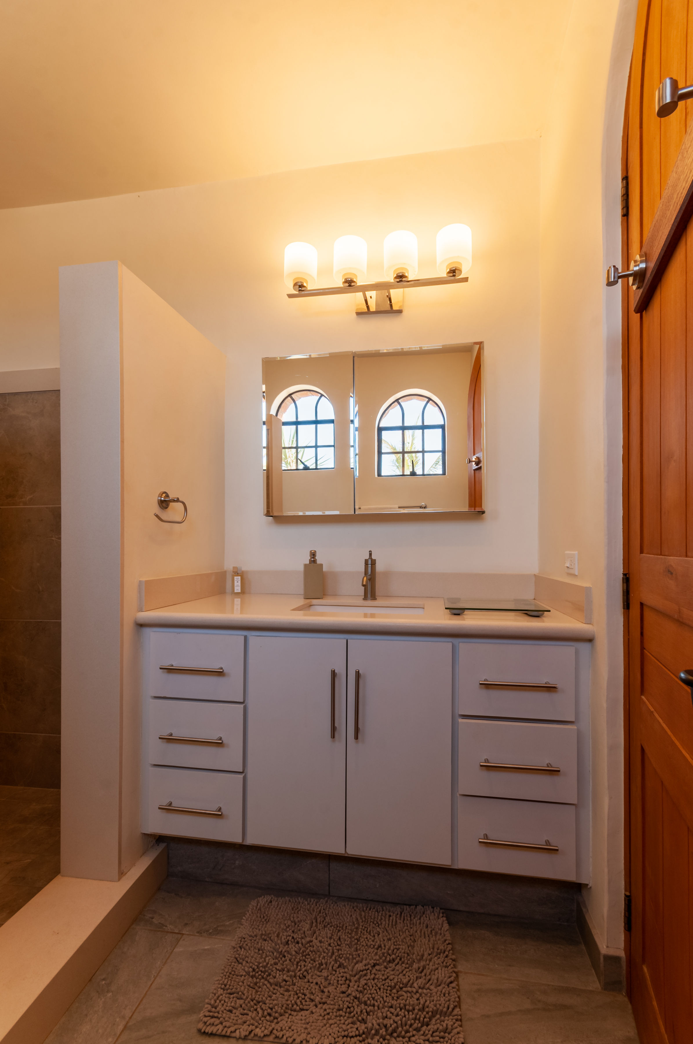 Bathroom in the adobe house with a white vanity, mirrored cabinet reflecting arched windows, and a tiled walk-in shower