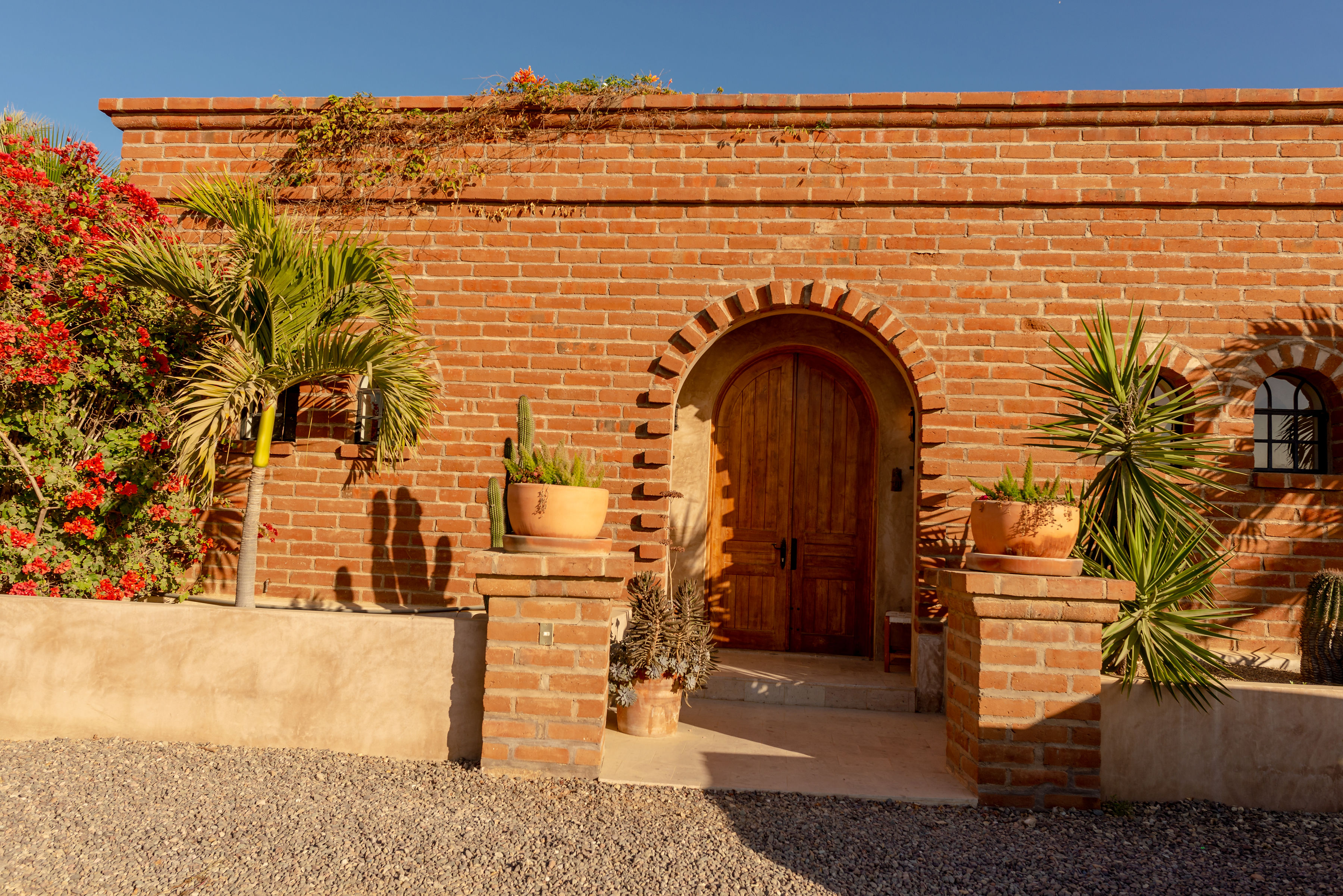 Brick facade of the adobe house at golden hour, with an arched wooden front door, potted cacti and bougainvillea