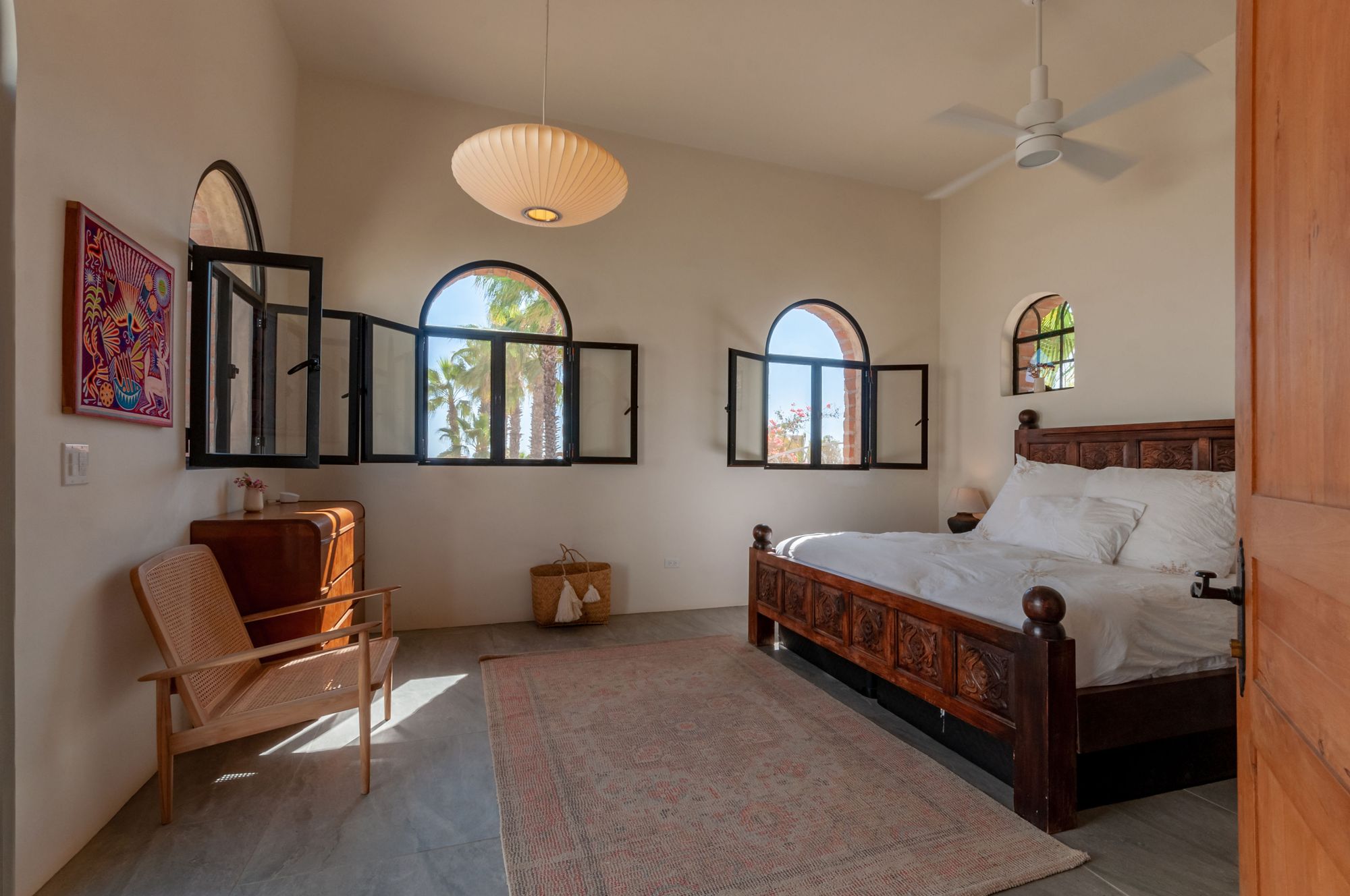 Bedroom with a carved wooden bed and three arched windows looking onto the palms