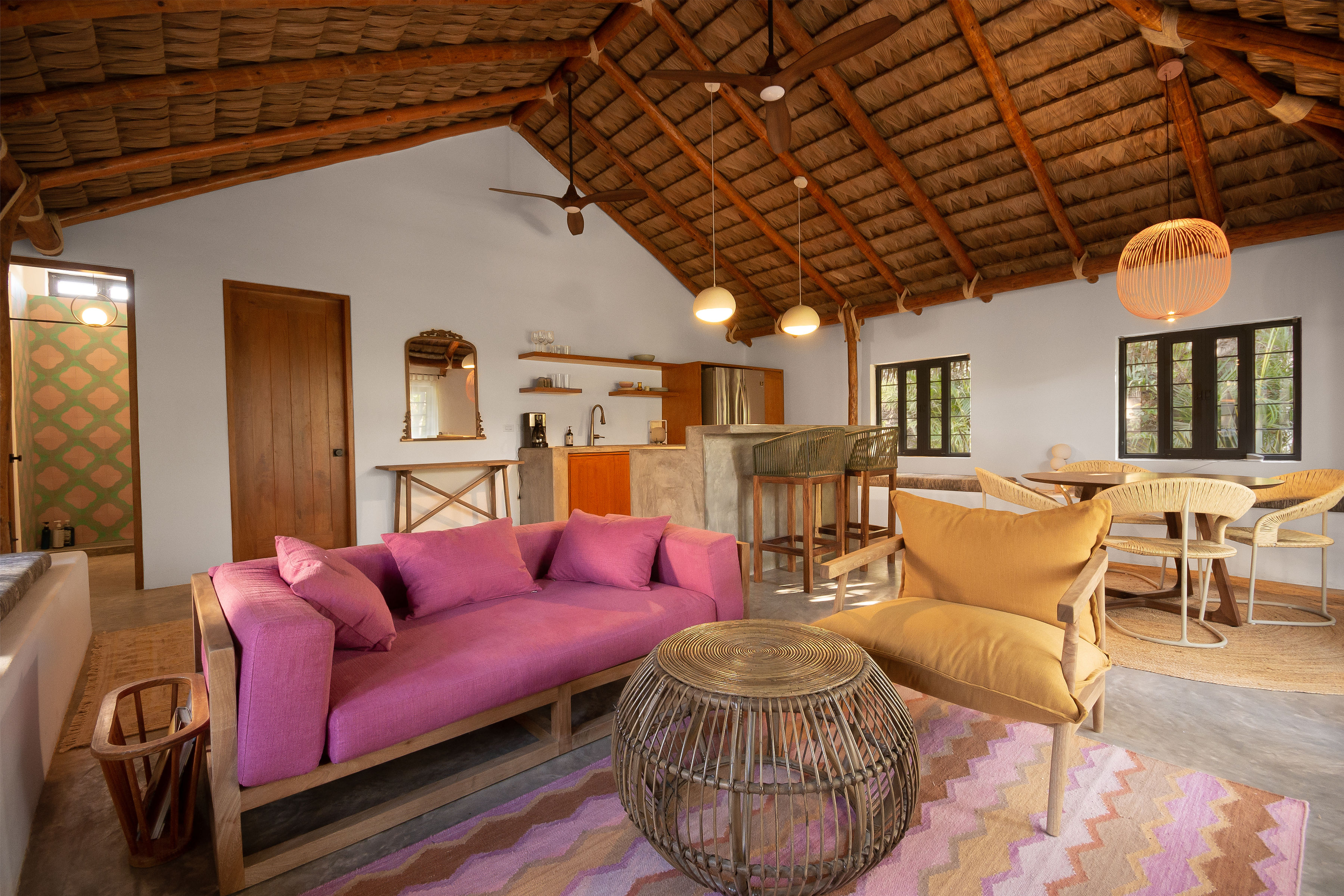 Wider view of the casita's living and dining area beneath the palapa roof, with the kitchen counter and bar stools beyond