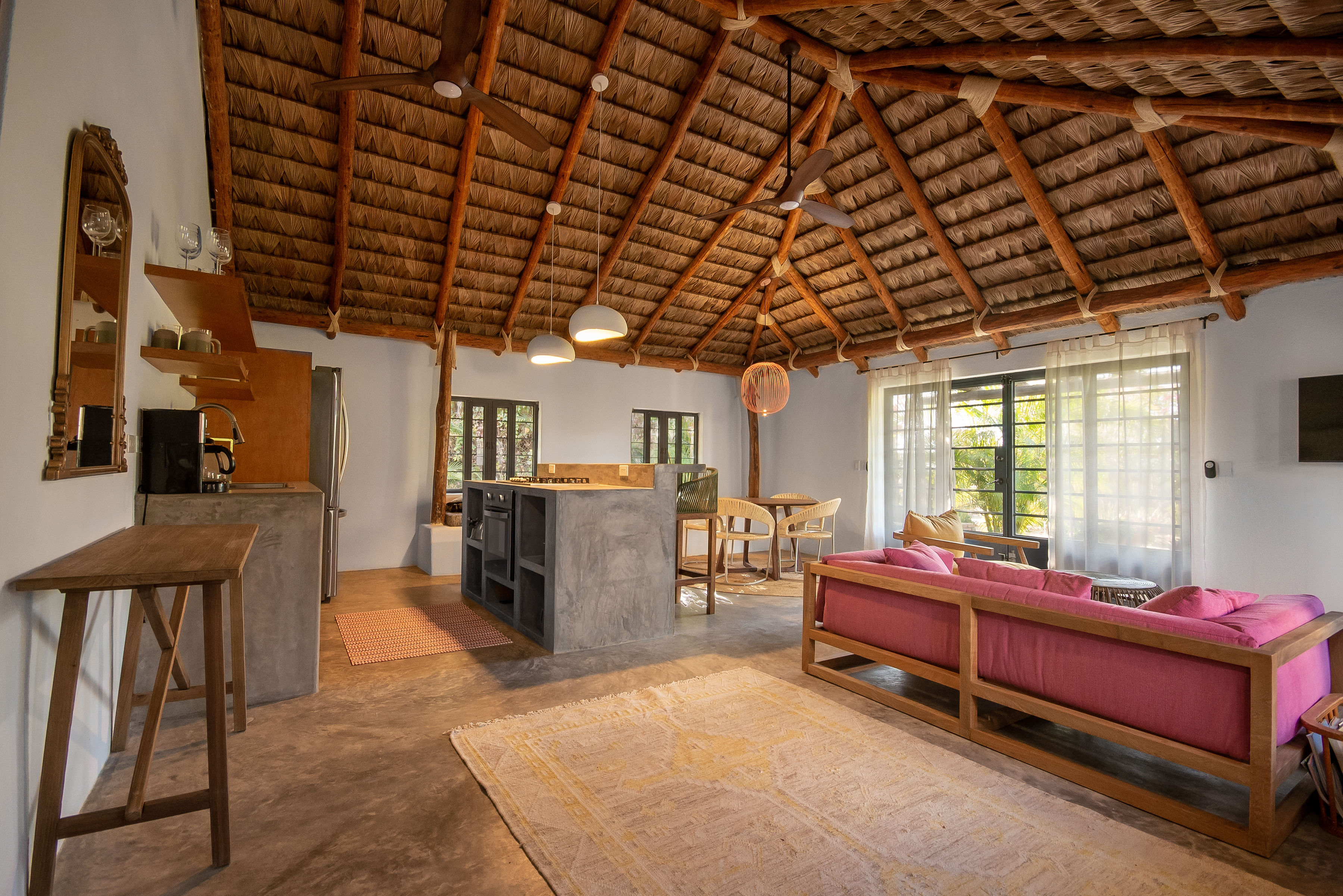 The casita's kitchen and living room under a palapa ceiling, with a concrete island, dining table and pink sofa