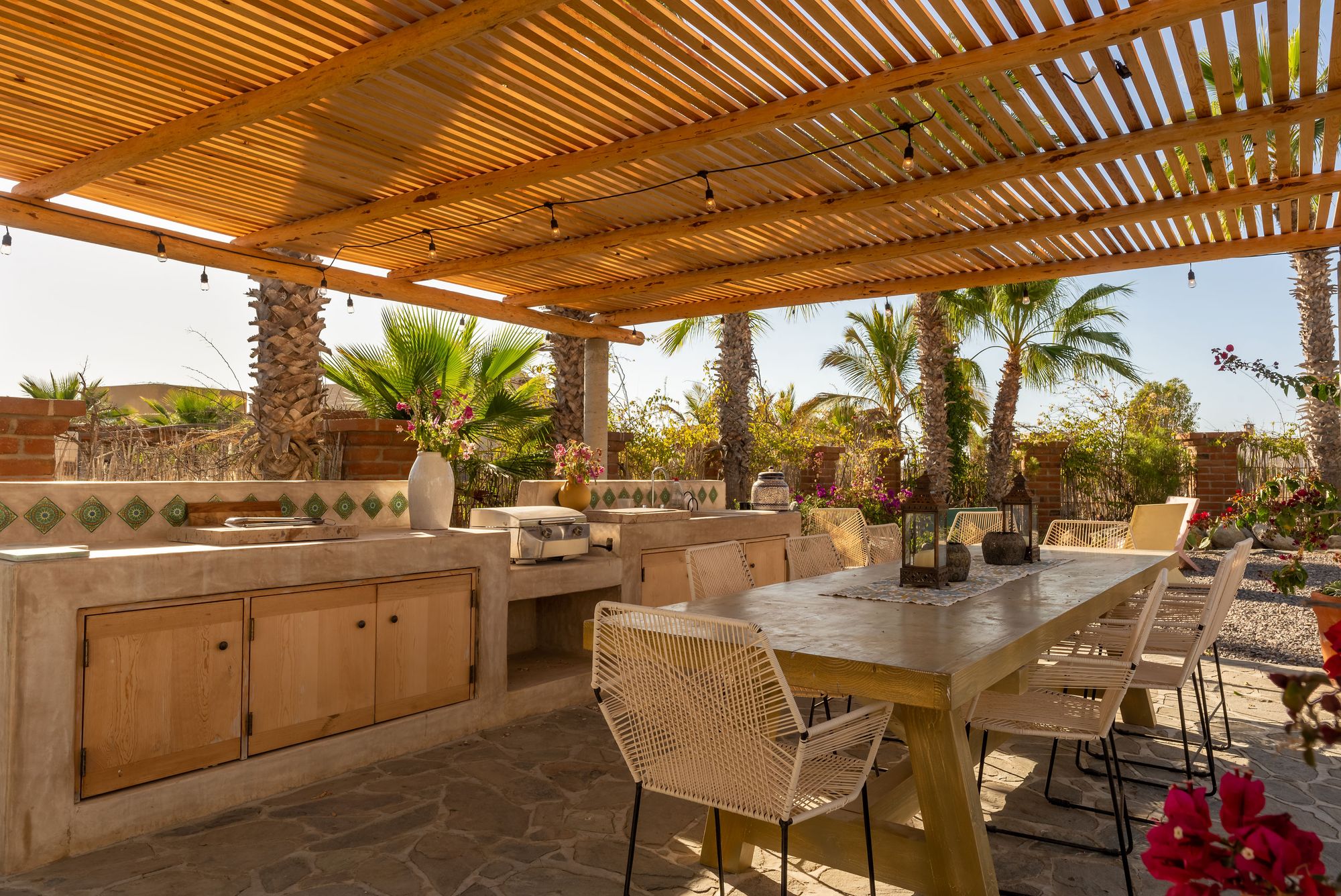 The long outdoor table under the wooden slat pergola beside the open-air kitchen