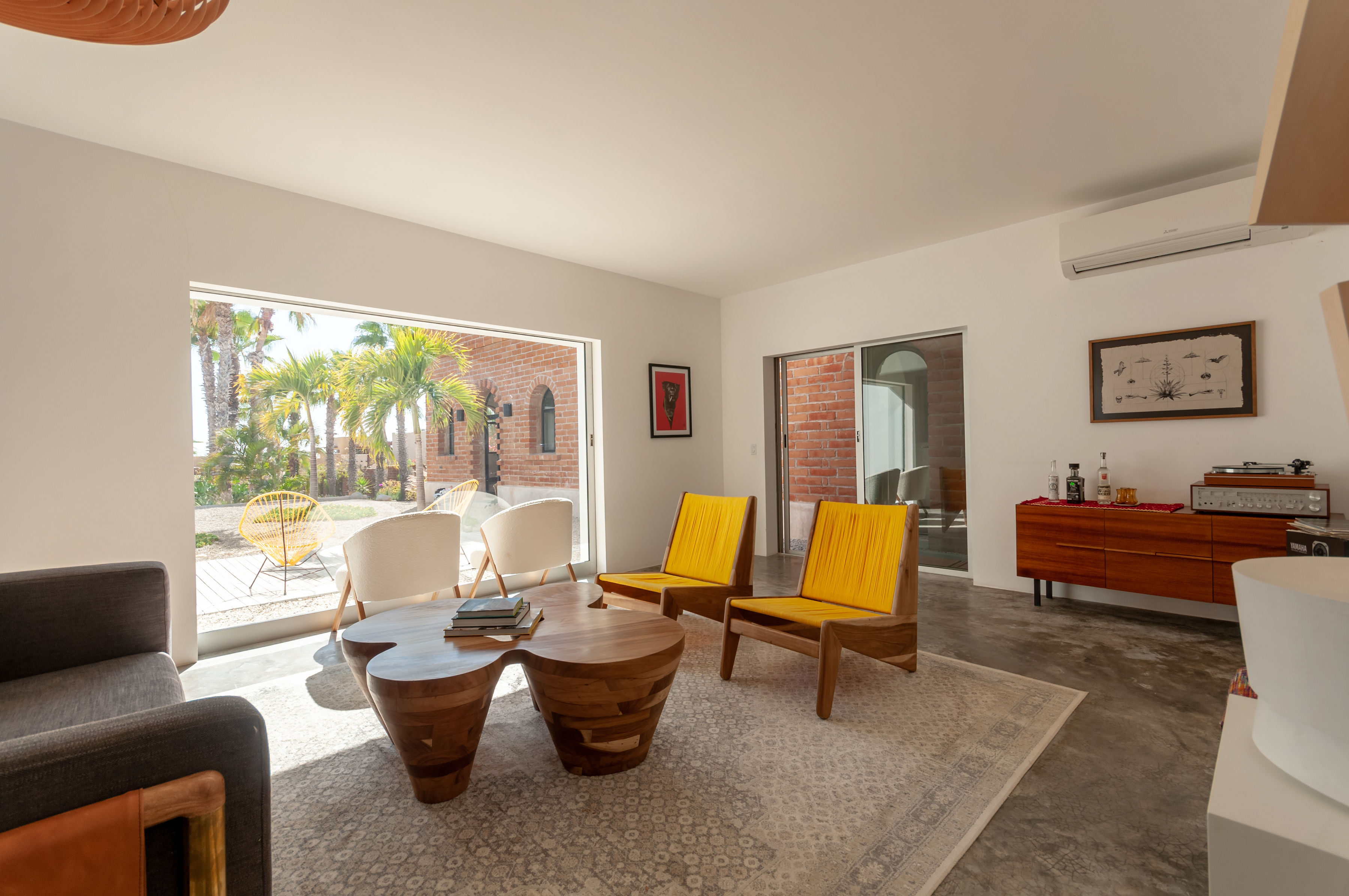 Shared living room with yellow woven lounge chairs, boucle armchairs, a sculpted wooden table and sliding doors open to the palms and brick courtyard