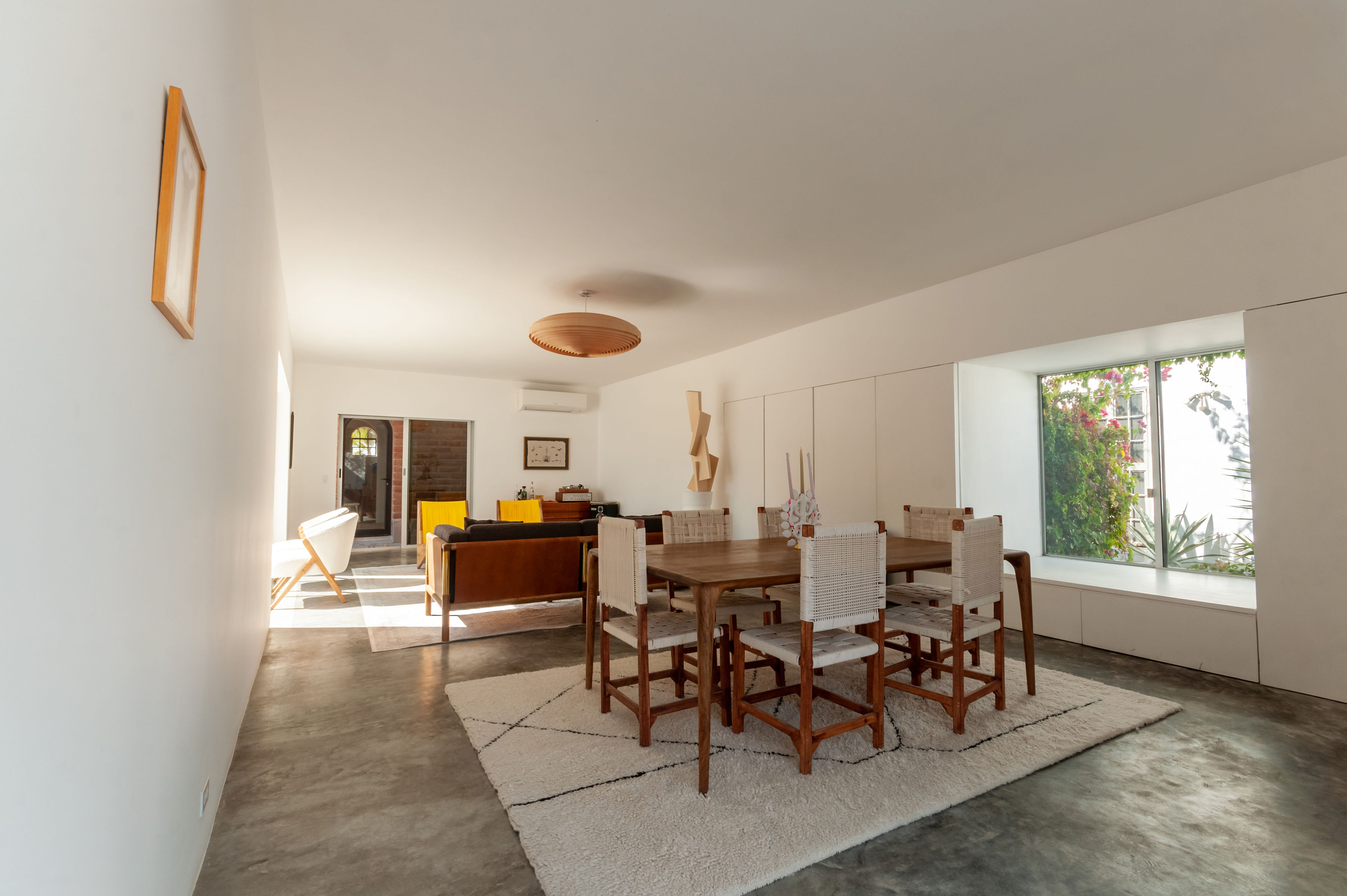 Dining table with woven chairs on a wool rug, looking through to the living room and the bougainvillea outside the window