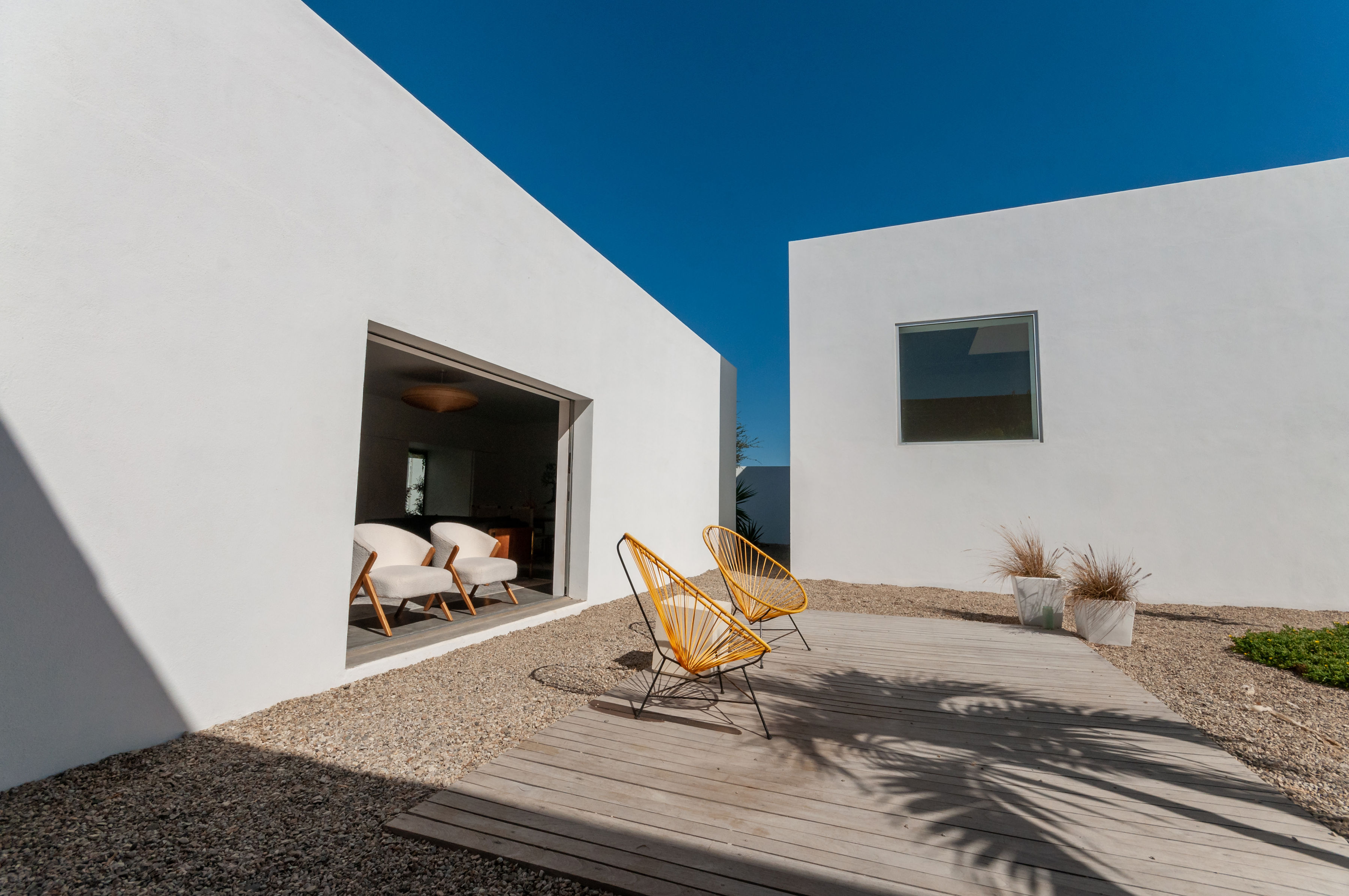 White stucco volumes of the studio building with a wide sliding opening, yellow Acapulco chairs on a timber deck and gravel garden