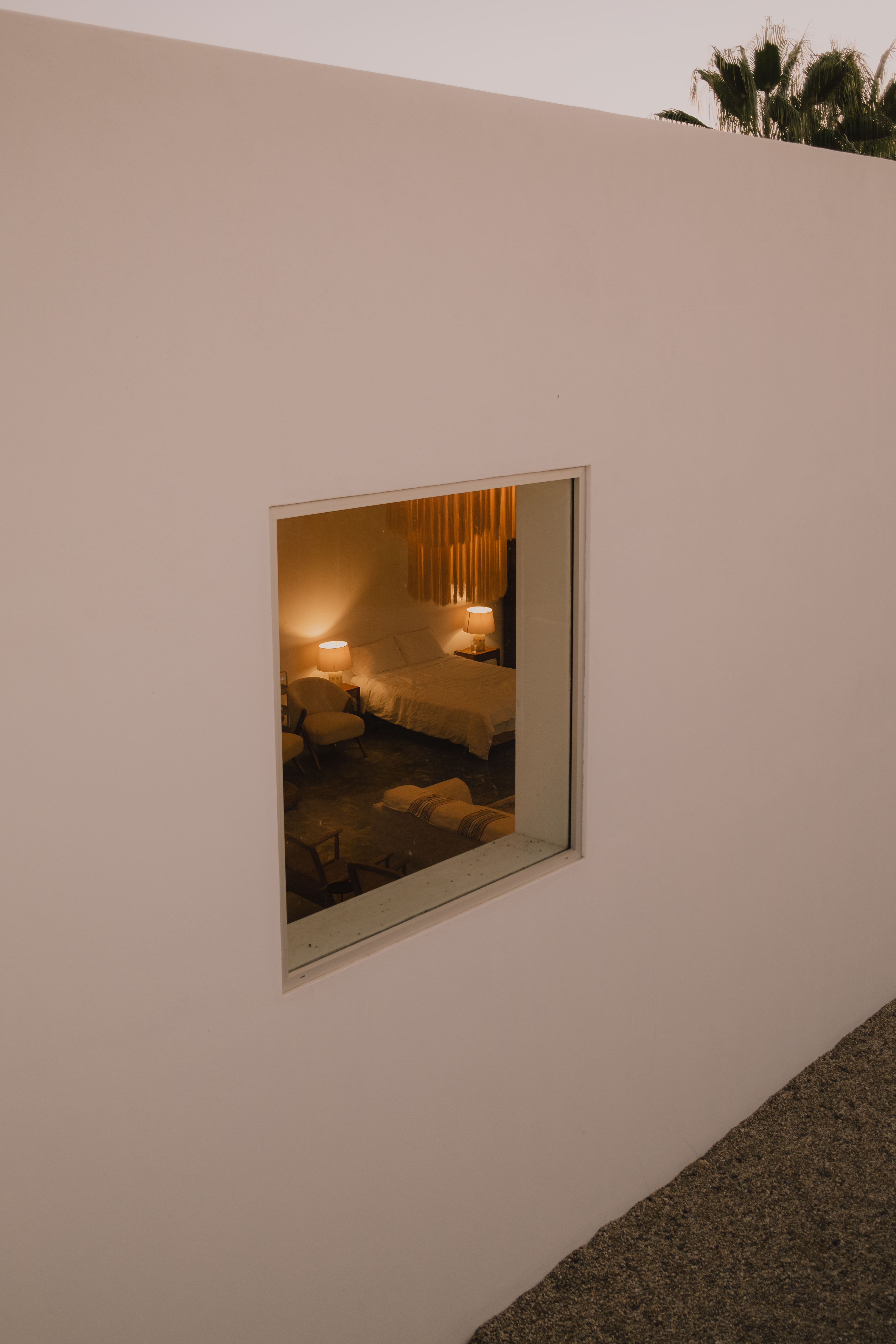 The studio's square window at twilight, lamplight showing the bed and seating inside the white stucco wall